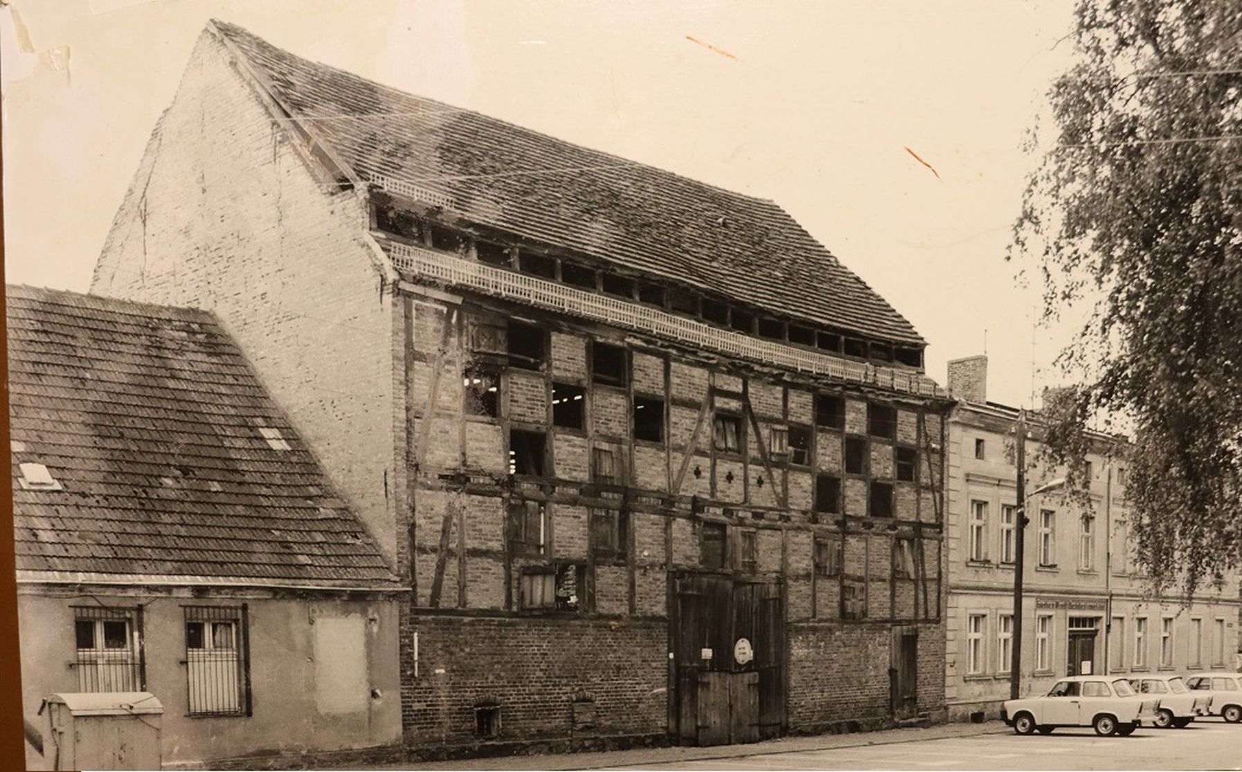 SW Foto. Eine Hausreihe von drei Gebäuden. In der Mitte steht eine große alte Tabakscheune mit Fachwerk und vielen Luken.