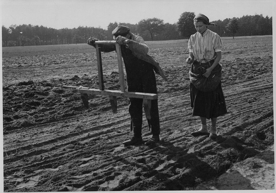 Ein Mann und eine Frau stehen auf einem Feld. Er hält einen Furchenzieher vor sicht, sie trägt eine Schürze mit Saatgut und verteilt dieses in den Furchen.