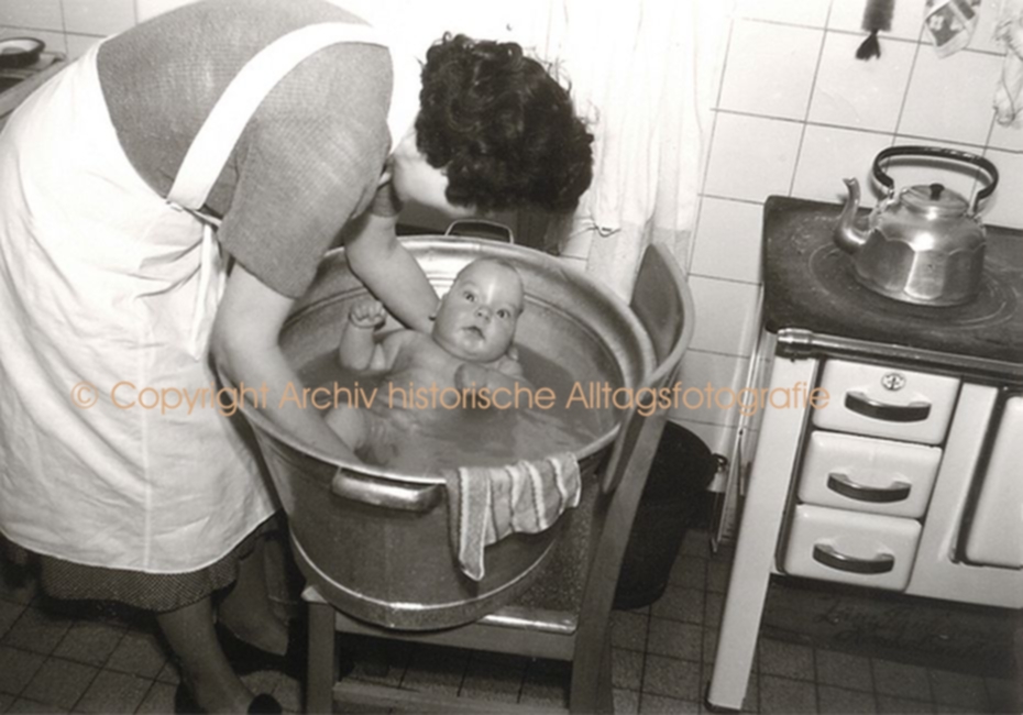 Ein Baby wind in einer Zinkbadewande, die in der Küche steht, von einer Frau gebadet.