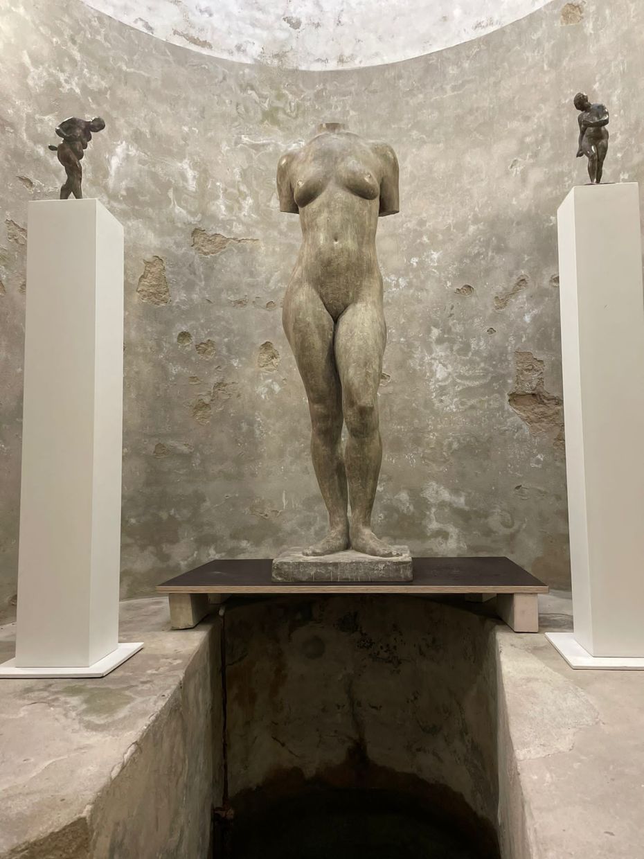 Über einem Wasserbecken steht eine Skulptur. Sie ist lebensgroß und zeigt einen Frauenkörper. Rechts und links daneben zwei weitere, kleine Skulpturen auf Sockeln.