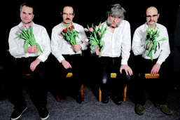 Foto: Die 4 Bandmitglieder sitzen mit weißen Hemden, schwarzen Hosen und jeweils einem Tulpenstrauß in einer Hand auf Holzstühlen in einer Reihe.