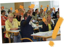 schräg gestelles Foto von einer Abstimmung in einer Stadtverordnetenversammlung mit gelbem Ausrufungszeichen