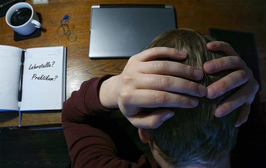 Foto: Blick von oben auf eine Tisch mit Laptop, Tasse und Kalender, darüber Hände am Kopf haltender Jugendliche