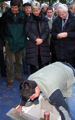 Foto: 3er-Gruppe blickt herab auf ein Bodenloch, in dem ein Bauarbeiter eine Kupferhülse einmauert.