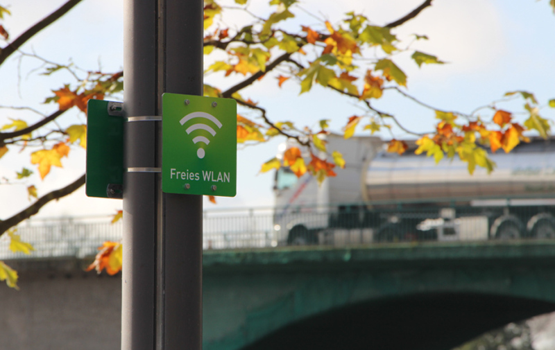 Foto: WLAN-Schild an der Laterne, im Hintergrund ist die Stadtbrücke mit Laster erkennbar